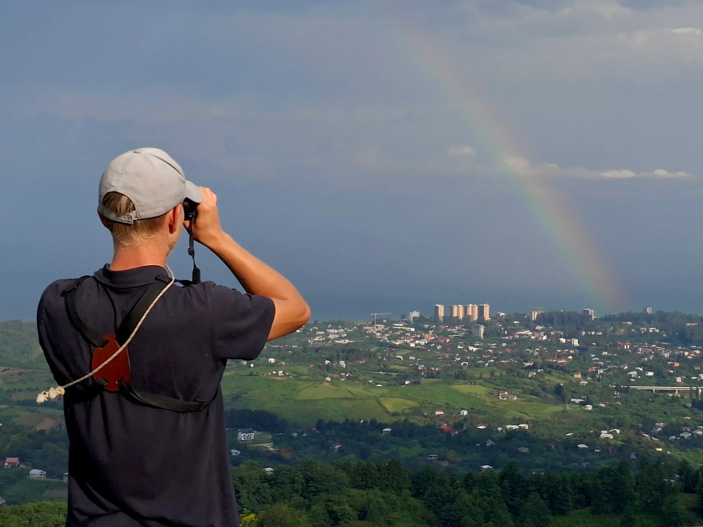 Viktor watching through binoculars with a rainbow over Batumi behind him.
